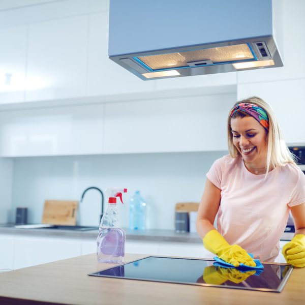 Worthy cheerful caucasian blond housewife with rubber gloves on hands cleaning stove. Kitchen interior.